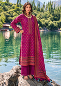 A woman stands barefoot on a rock by a lake wearing a magenta three-piece ethnic outfit with gold embroidery, a matching dupatta, and gold jewelry, with a scenic mountain and lake background.