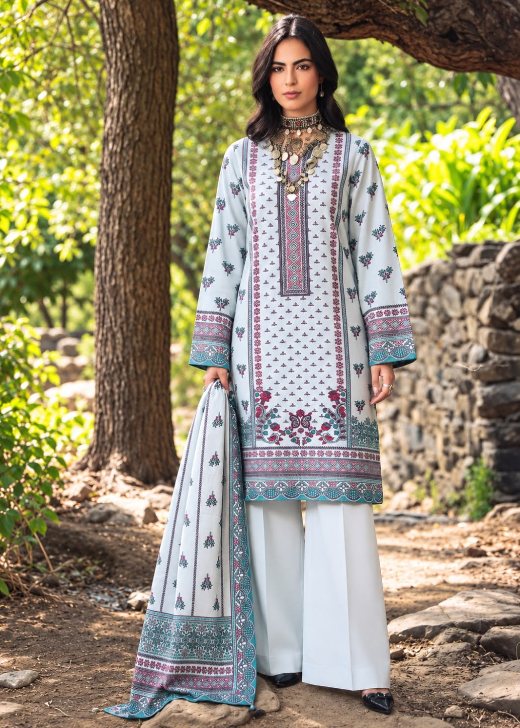 A woman stands outdoors in a wooded setting wearing a traditional three-piece salwar kameez outfit. The outfit consists of a white long-sleeve kurta with blue, purple, and red floral embroidery and geometric patterns, paired with white wide-leg trousers and a matching white dupatta with a blue and purple border.