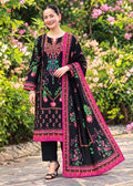 A woman stands outdoors in a garden wearing a black traditional outfit with bright pink borders and colorful floral embroidery, paired with black pants and black heels. The background features lush green foliage and pink flowers.