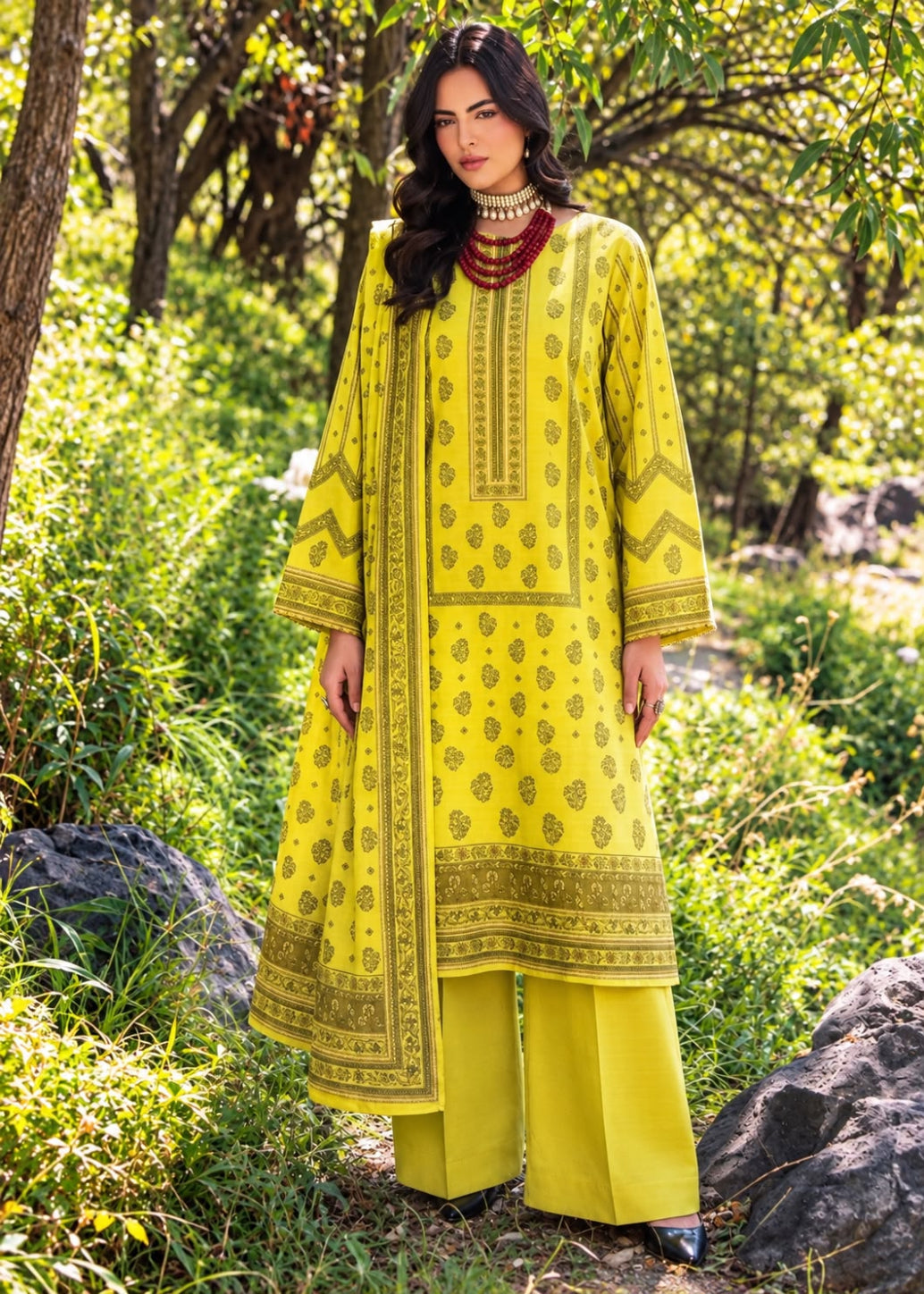 A woman wearing a bright yellow traditional three-piece kurta set with gold embroidery, standing outdoors in a lush green garden setting.