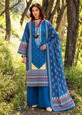 A woman stands outdoors in a wooded area wearing a traditional blue three-piece salwar kameez suit with colorful floral prints and embroidery, matching wide-leg trousers, and a draped dupatta.
