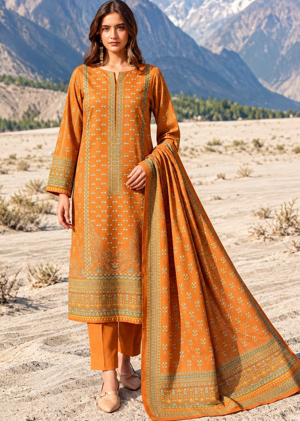 A woman stands outdoors in a desert landscape wearing a traditional three-piece outfit with an orange kurta, matching pants, and a draped dupatta, all decorated with green and beige geometric patterns. Mountains are visible in the background.