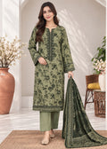 A woman models a three-piece olive green ethnic suit with black floral embroidery, matching trousers, and a coordinating dupatta in a bright, modern room setting.