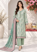 A woman models a traditional three-piece salwar kameez suit in mint green with intricate floral prints, paired with matching green trousers and a printed dupatta, set against a bright, modern indoor backdrop.