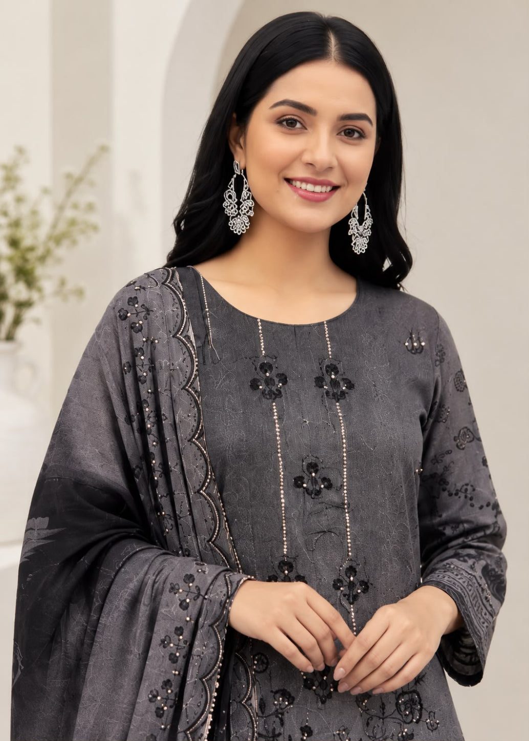 A woman modeling a dark gray three-piece salwar kameez suit with black floral embroidery and beadwork, paired with matching embroidered dupatta and silver earrings.