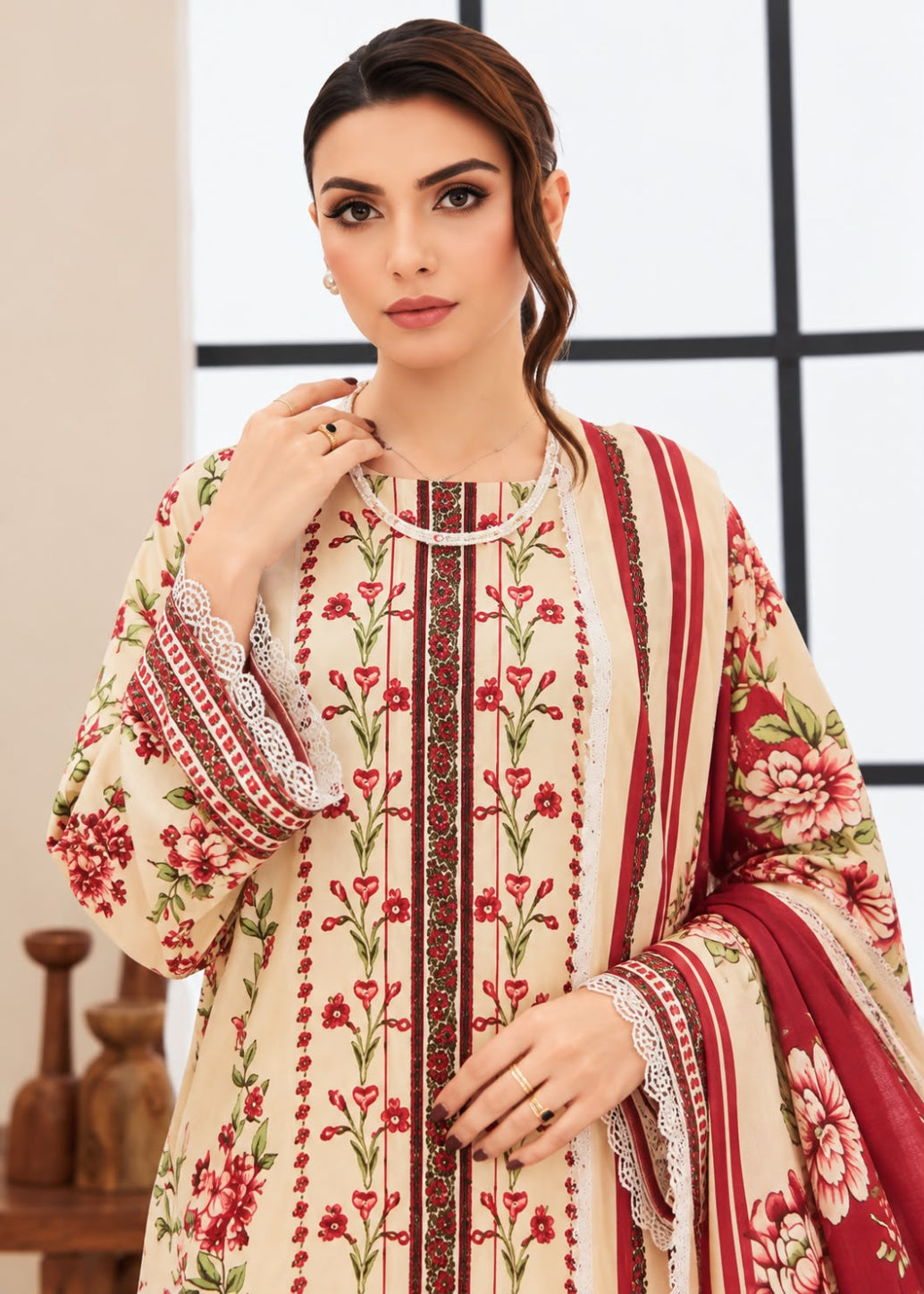 A woman modeling a traditional embroidered kurta set in cream with red and green floral patterns, paired with a matching red dupatta, set against a modern interior background.