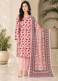 A woman models a three-piece pink ethnic suit set featuring a floral printed kurta, solid pink trousers, and a matching printed dupatta with a geometric border, set against an elegant indoor background with potted plants.