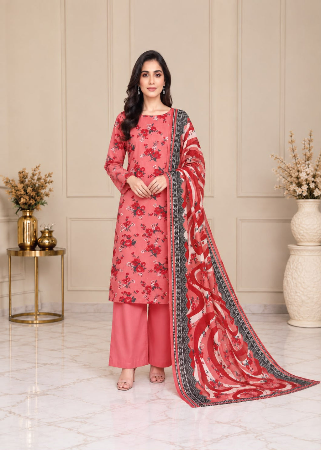 A woman models a traditional Indian salwar kameez suit set in a decorated indoor setting. The set features a coral pink floral printed kameez, matching pink trousers, and a red and pink patterned dupatta with a black border.