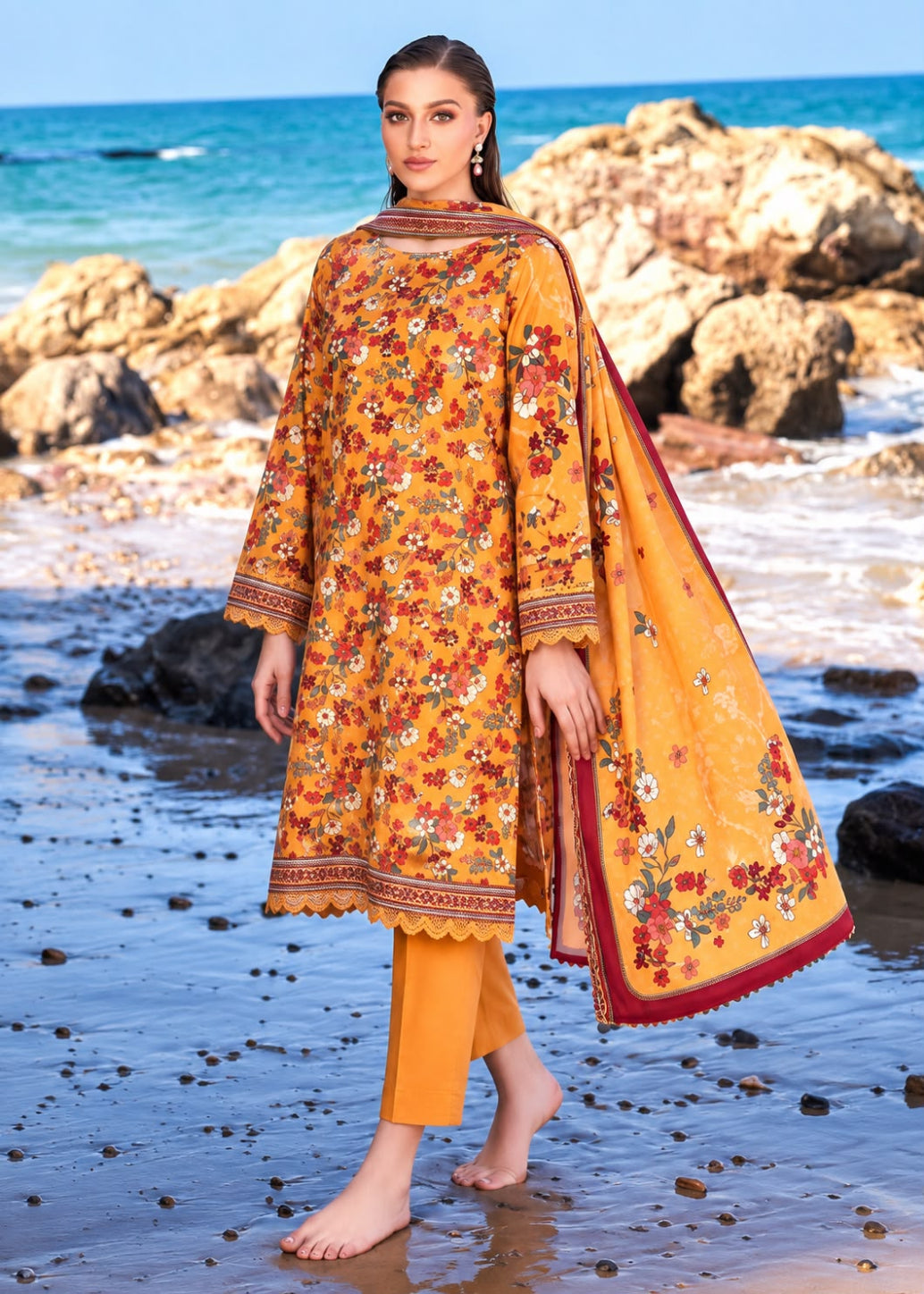 A woman stands barefoot on a beach wearing a bright yellow embroidered kurta set with matching pants and a draped dupatta. The outfit features dense floral embroidery in red, orange, and white, with a deep red border.