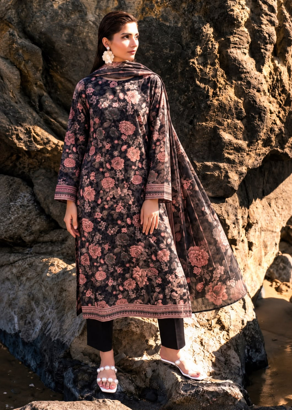 A woman stands outdoors against large rock formations wearing a dark floral salwar kameez outfit with a matching dupatta. The outfit has a black base with pink and rose floral patterns and patterned borders, paired with black pants and white slide sandals.