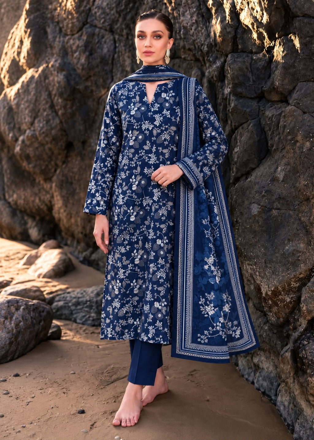 A woman stands barefoot on a sandy shore against a large rock wall, wearing a navy blue floral printed kurta with matching navy pants and a navy dupatta featuring white border and floral embroidery.