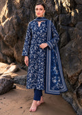 A woman stands barefoot on a sandy shore against a large rock wall, wearing a navy blue floral printed kurta with matching navy pants and a navy dupatta featuring white border and floral embroidery.