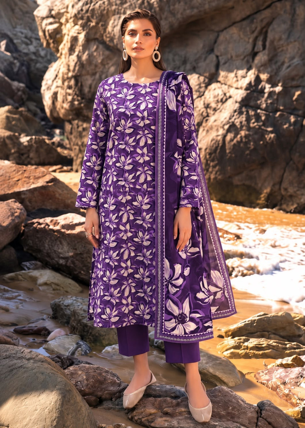 A woman stands outdoors on rocky shoreline rocks wearing a purple three-piece traditional outfit with white floral print and a matching dupatta.