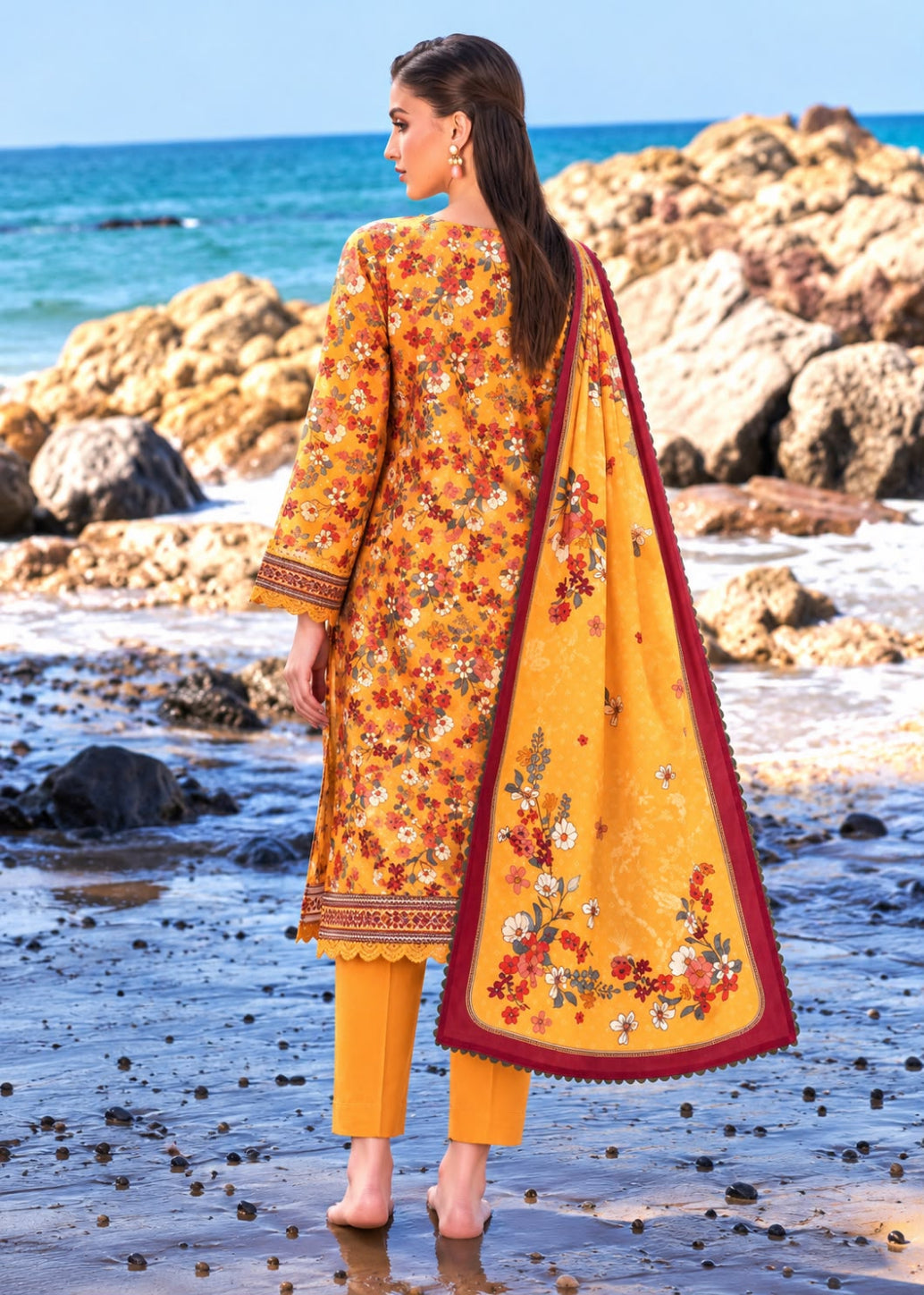 A woman stands on a rocky beach facing away from the camera, wearing a bright yellow traditional outfit with floral embroidery and a matching dupatta.