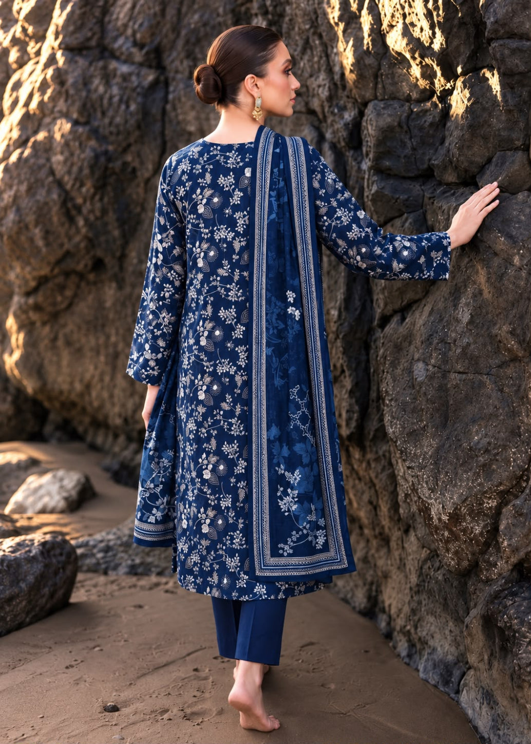 A woman stands barefoot against a rocky outdoor backdrop wearing a navy blue traditional outfit with white floral embroidery, matching pants, and a draped scarf.