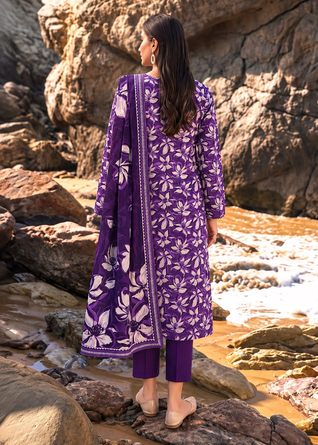 A woman stands on rocks by a rocky stream wearing a purple three-piece traditional outfit with a white floral print and matching dupatta.
