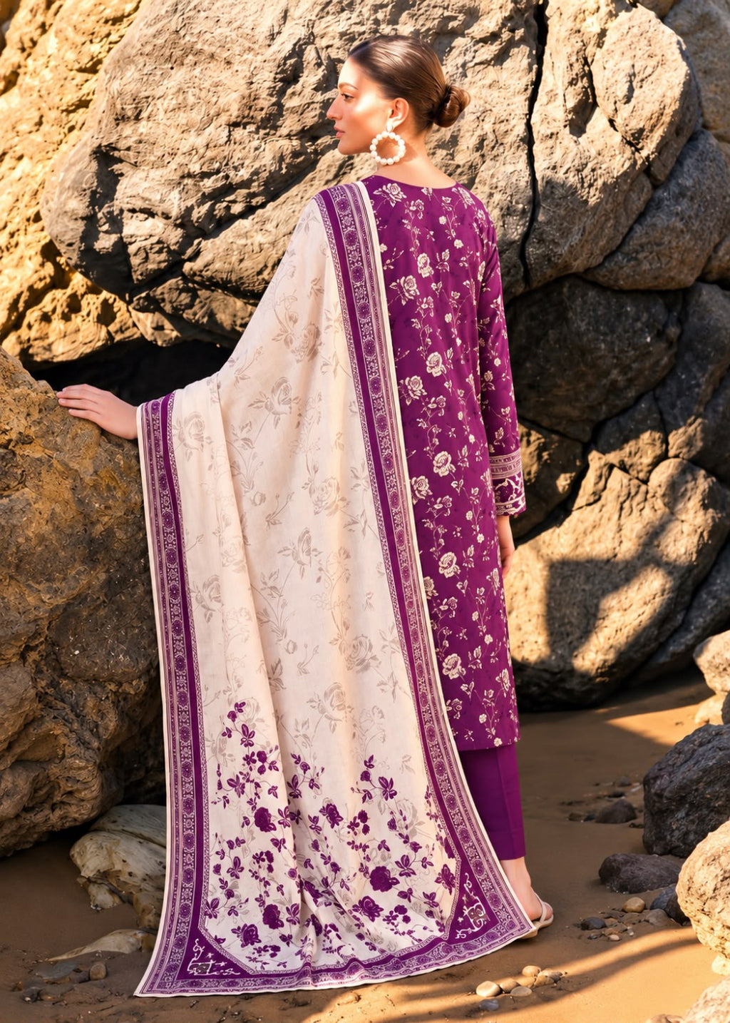 A woman stands outdoors against large rocks wearing a traditional outfit with a purple floral kurta and matching pants, paired with a cream dupatta featuring purple floral motifs and borders.