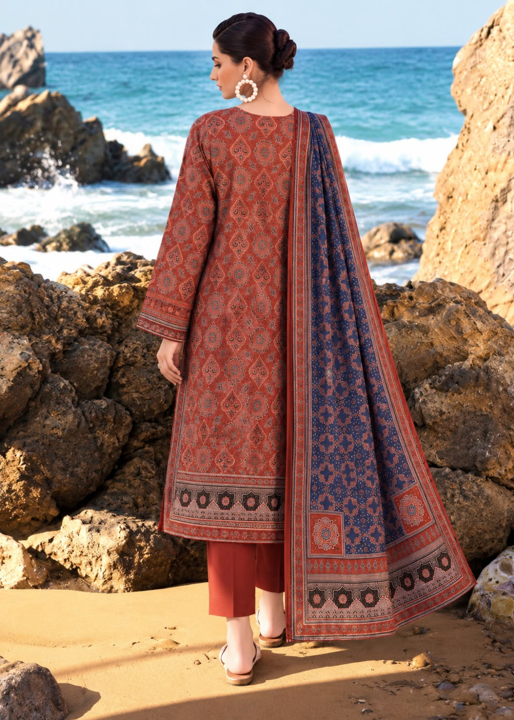 A woman stands on a sandy beach with large rocks and the ocean in the background, wearing a traditional three-piece outfit. The outfit consists of a rust-red printed kurta, matching pants, and a navy blue printed dupatta with red borders.