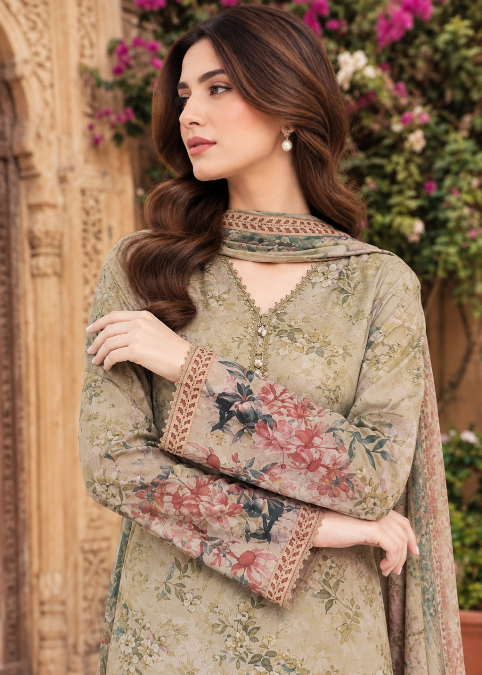A woman wearing a beige printed traditional outfit with floral embroidery and a matching dupatta, posed outdoors in front of a carved wooden door and greenery.
