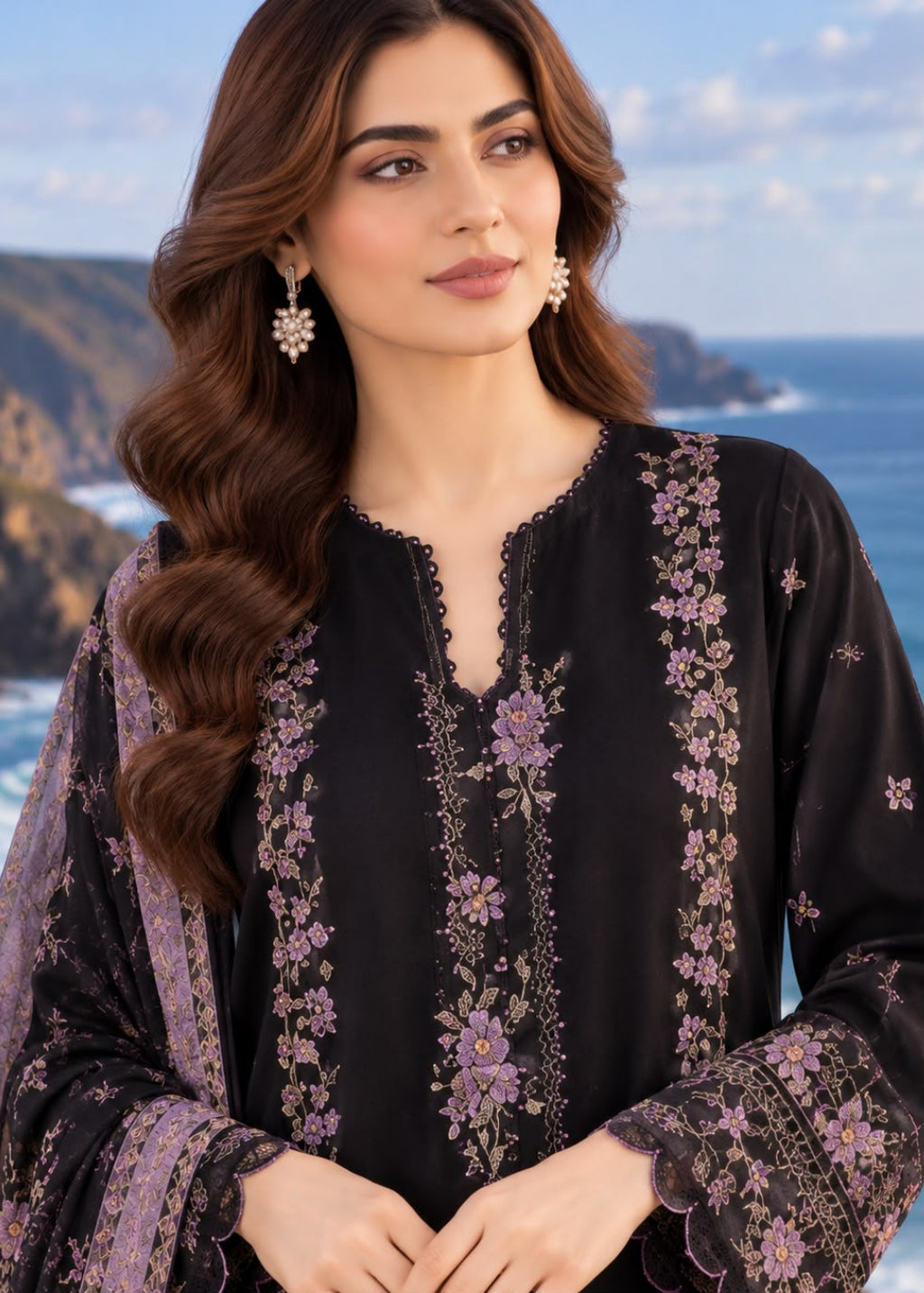 A woman wearing a black embroidered kurta with purple floral patterns and a matching sheer dupatta, posed against a scenic coastal background.