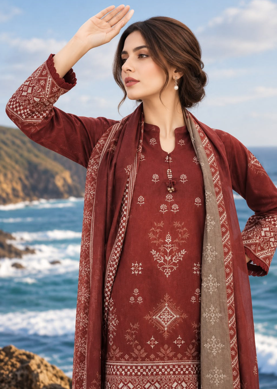 A woman wearing a rust-red printed ethnic kurta with a matching dupatta and a beige patterned outer layer, standing outdoors by the ocean with cliffs and waves in the background.