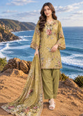 A woman stands on a rocky coastal cliff wearing a three-piece traditional outfit with a floral print. The set includes a mustard yellow kurta with pink and green floral embroidery, olive green pants, and a matching dupatta.