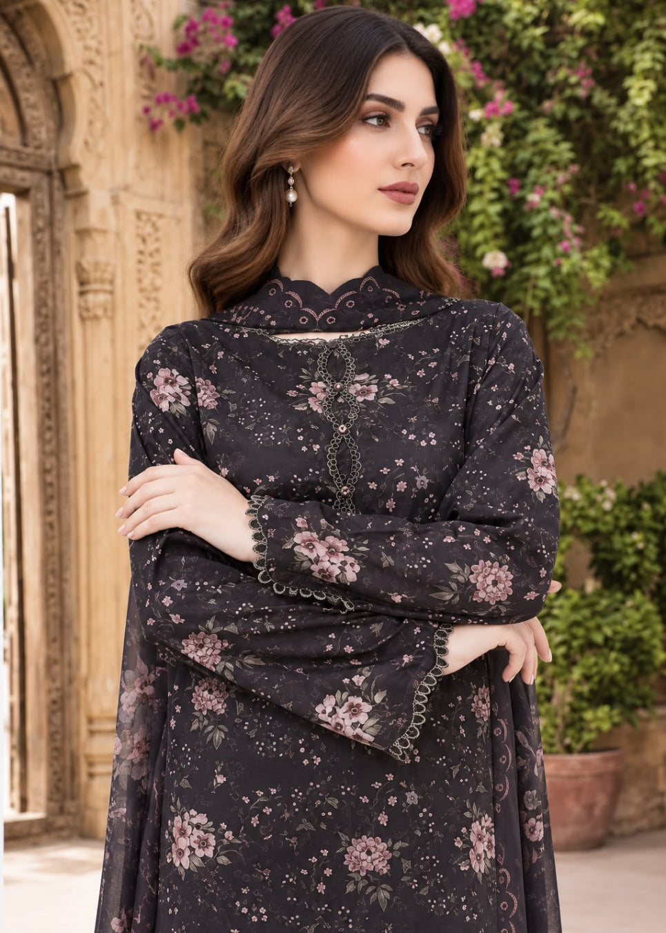 A woman wearing a dark charcoal black long-sleeve kurta with pink floral prints and scalloped lace trim, posed outdoors in front of a stone wall and greenery.