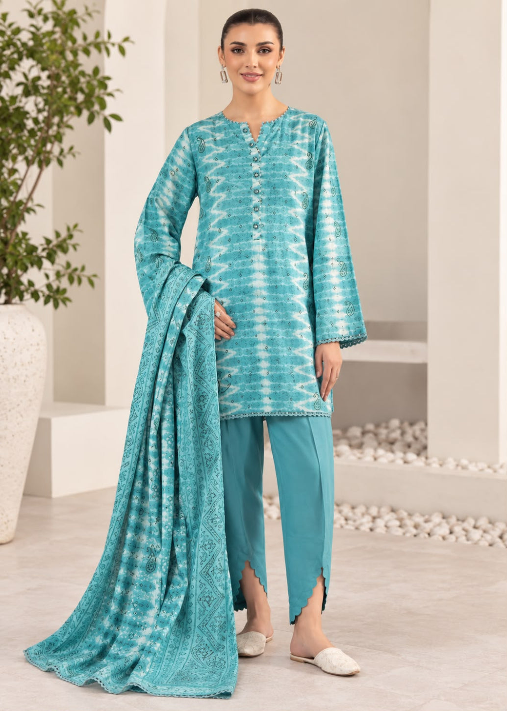 A woman modeling a teal blue and white printed salwar kameez suit with matching pants and a dupatta, standing in a modern indoor setting.