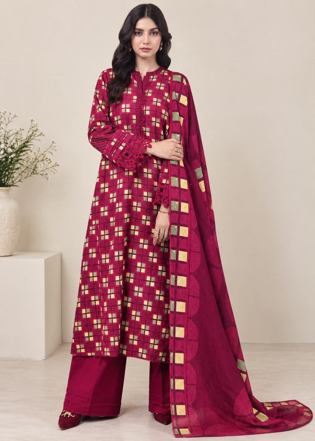 A woman models a traditional three-piece salwar kameez suit in deep magenta with a gold geometric checkered pattern, paired with matching wide-leg trousers and a long dupatta, set against a neutral beige background.