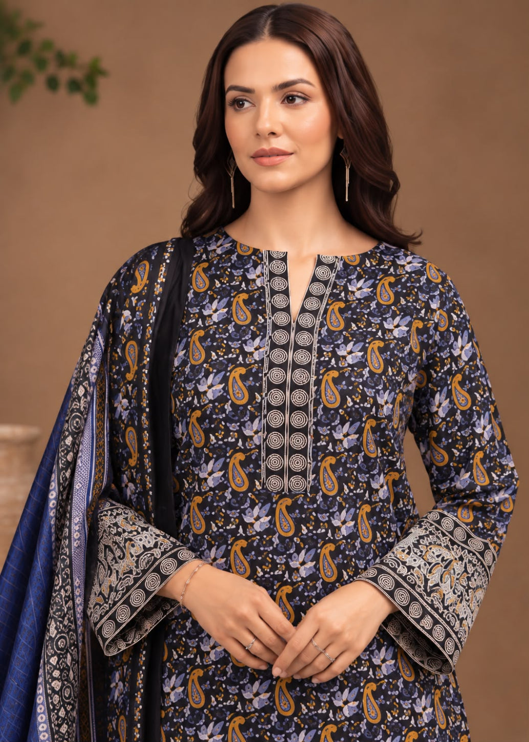 A woman modeling a navy blue printed salwar kameez suit with gold and gray paisley motifs, featuring a matching navy dupatta with gold and gray borders, set against a warm brown studio background.