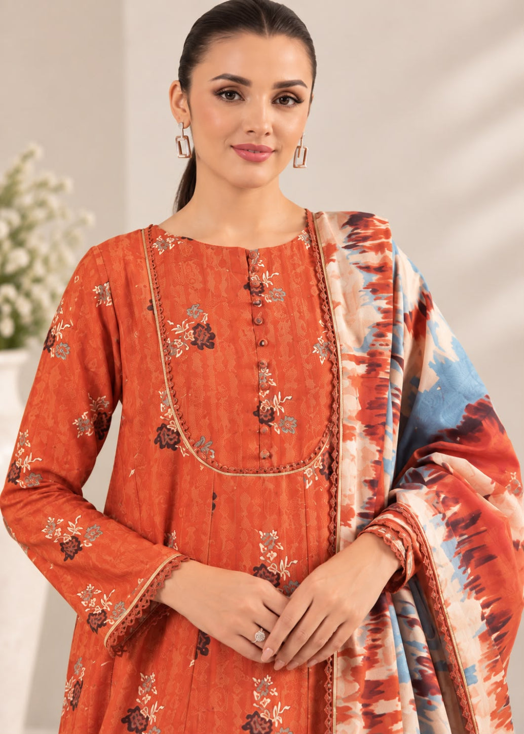 A woman modeling a rust-orange printed salwar kameez suit with a matching dupatta. The kurta features a floral print and decorative neckline, and the dupatta has a blue, orange, and white tie-dye pattern.