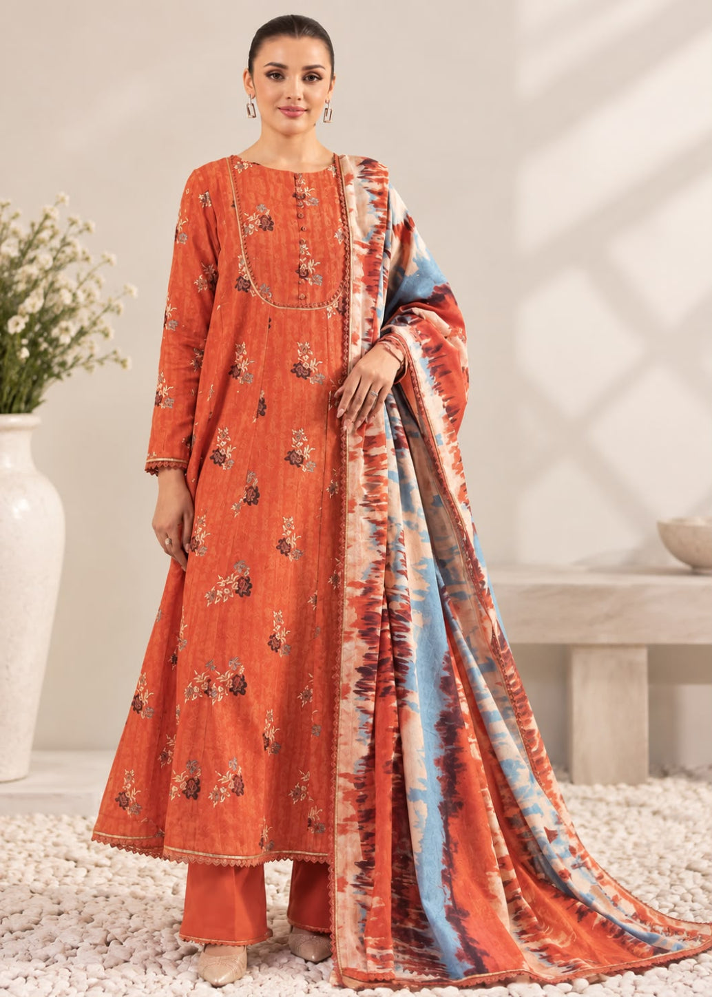 A woman models a traditional Indian salwar kameez suit set in terracotta orange, featuring a floral print, matching trousers, and a coordinating dupatta with a blue and orange tie-dye pattern, set against a neutral indoor background.