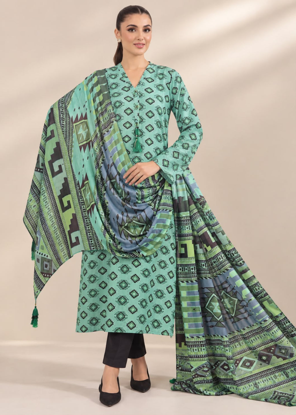 A woman models a mint green three-piece salwar kameez suit with geometric patterns, paired with black pants and black heels, against a beige studio background.