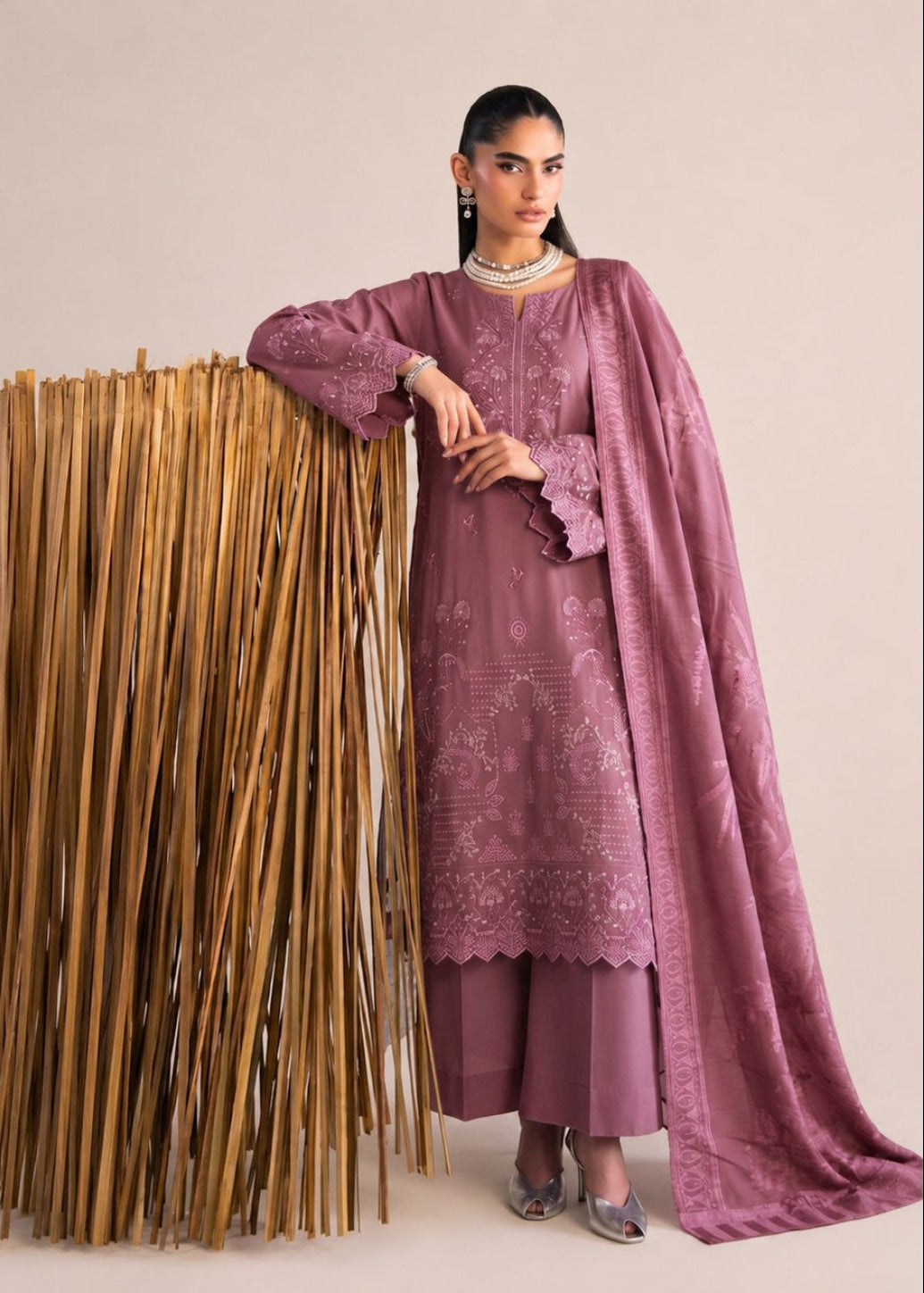 A woman models a traditional three-piece salwar kameez suit in dusty mauve with intricate silver embroidery, standing next to a large decorative stand of vertical wooden sticks against a solid beige studio background.