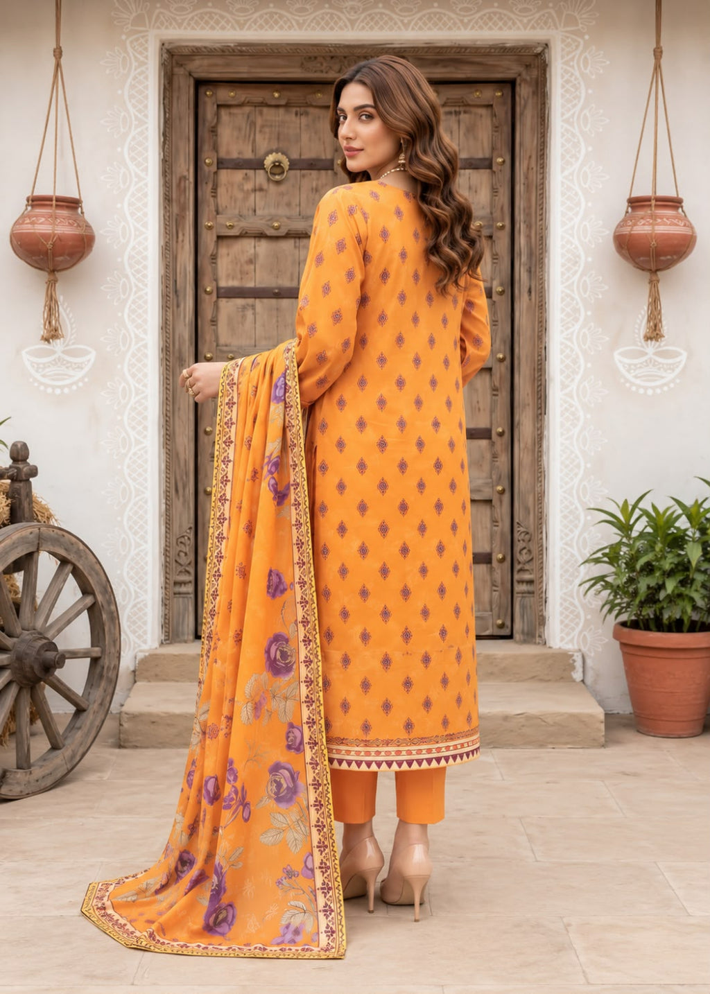 A woman stands in front of a rustic wooden door wearing a bright orange traditional outfit with small dark geometric motifs and a matching orange dupatta featuring purple floral prints and gold border detailing.
