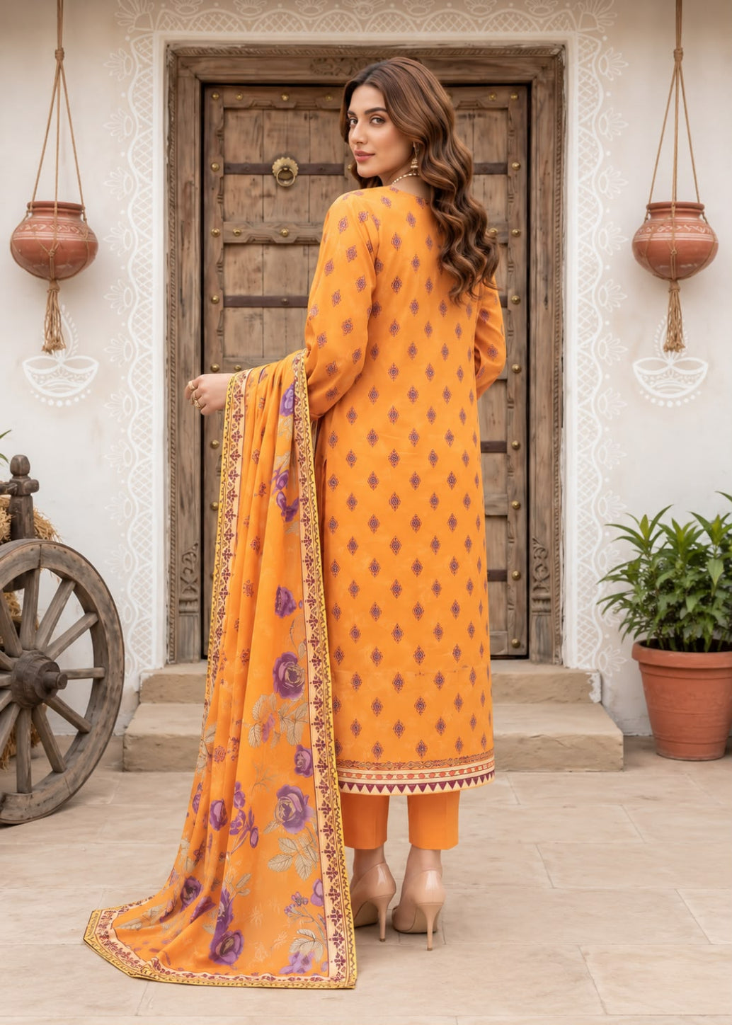 A woman stands in front of a rustic wooden door wearing a bright orange traditional outfit with small dark geometric motifs and a matching orange dupatta featuring purple floral prints and gold border detailing.