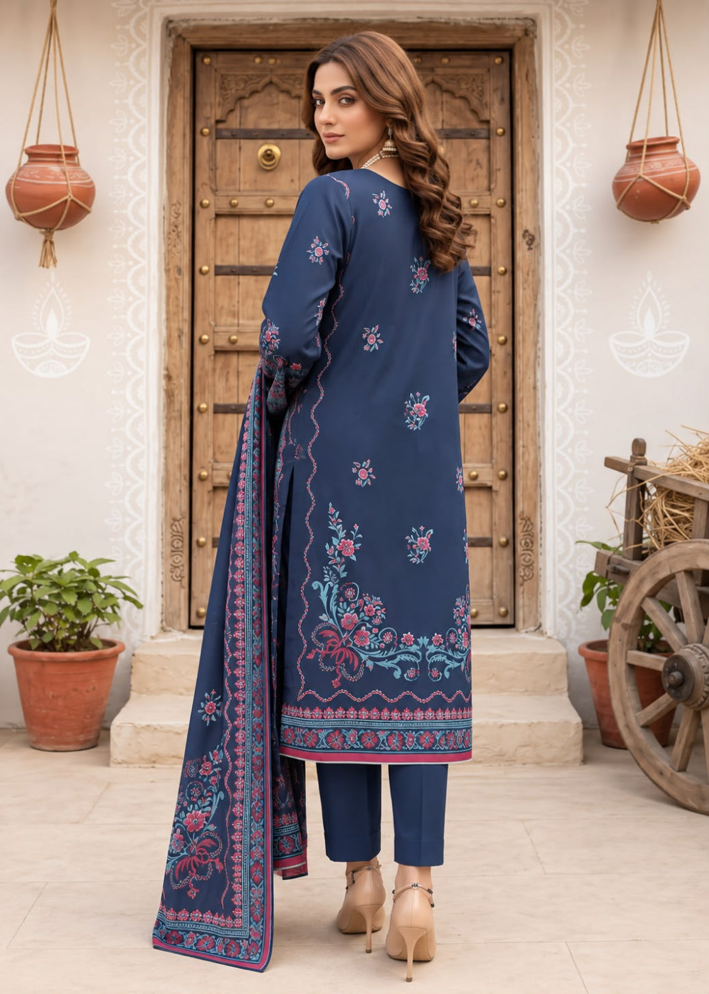 A woman stands with her back to the camera wearing a navy blue three-piece salwar kameez suit with pink and green floral embroidery, paired with matching navy trousers and a navy dupatta with a pink border, set against a rustic wooden door background.