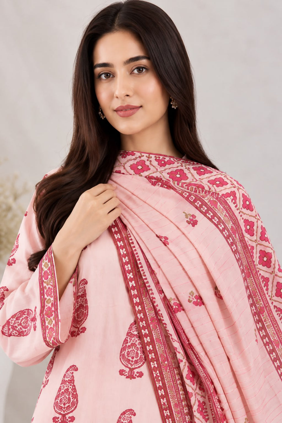 A woman modeling a light pink traditional outfit with a matching patterned dupatta. The outfit features dark pink paisley and floral motifs, and the draped scarf has a geometric border design, set against a soft neutral background.