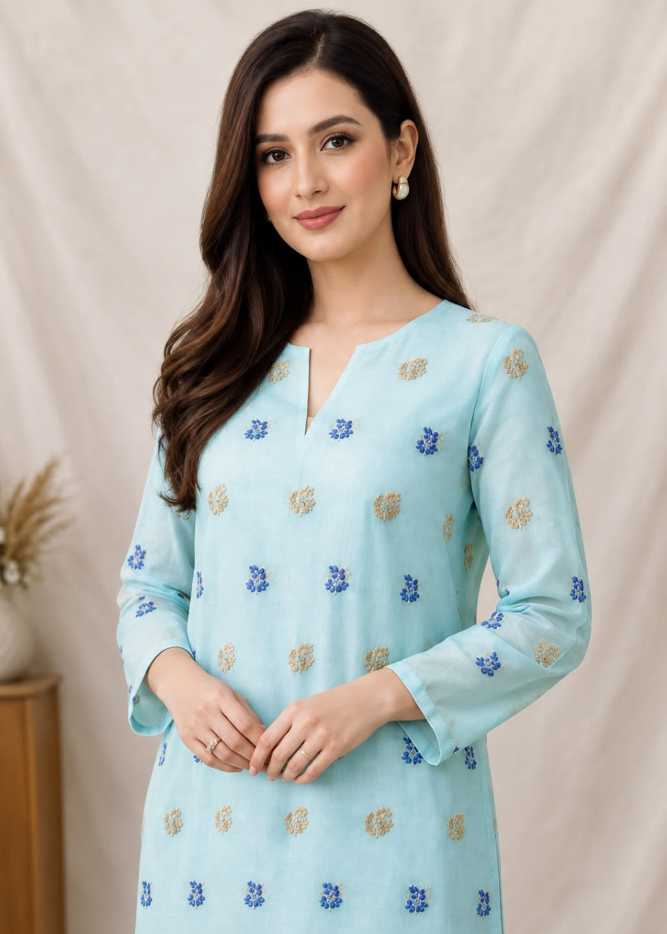 A woman modeling a light blue three-quarter sleeve kurta with small blue and gold floral prints, set against a neutral beige fabric backdrop.