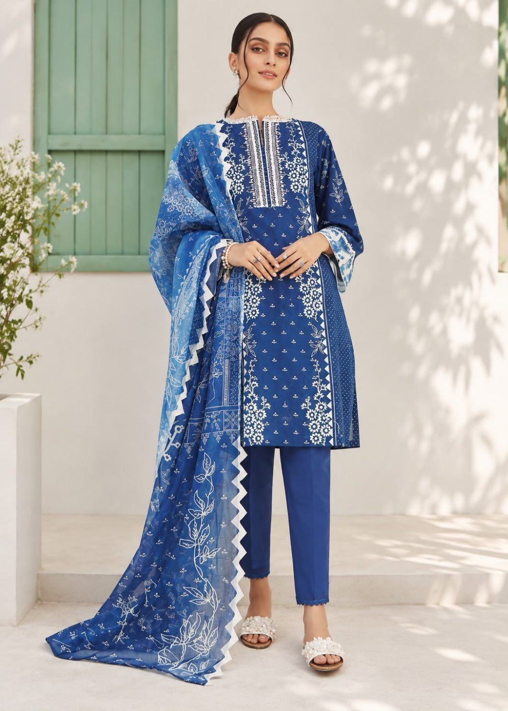 A woman models a three-piece blue ethnic suit set featuring a printed kurta, matching trousers, and a sheer printed dupatta with white borders, set against a white wall with green shutters.