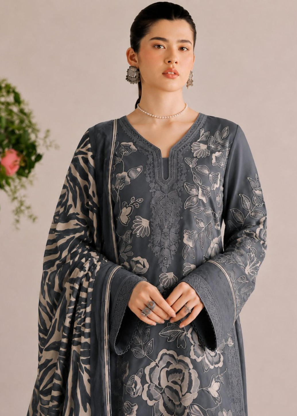A woman models a dark gray three-piece ethnic suit with intricate silver floral embroidery, featuring a V-neck kurta, matching wide-leg trousers, and a coordinating dupatta, set against a soft beige background.