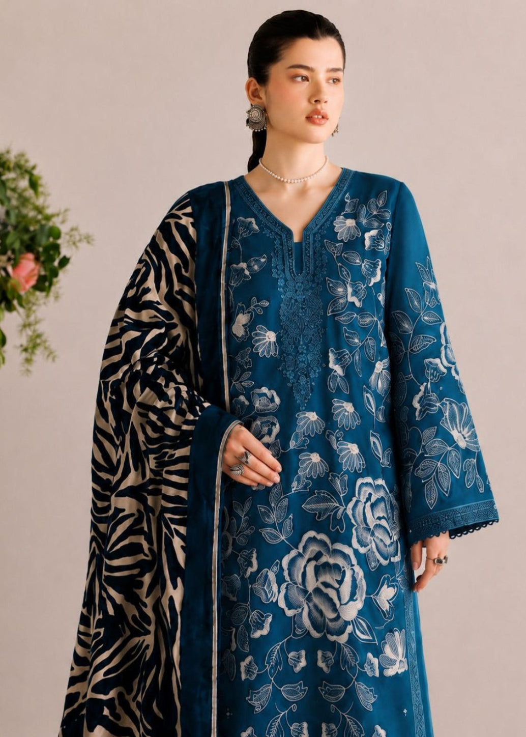 A woman models a traditional three-piece salwar kameez suit in deep teal blue. The kurta features intricate silver floral embroidery, and she pairs it with a navy and beige zebra-print dupatta.
