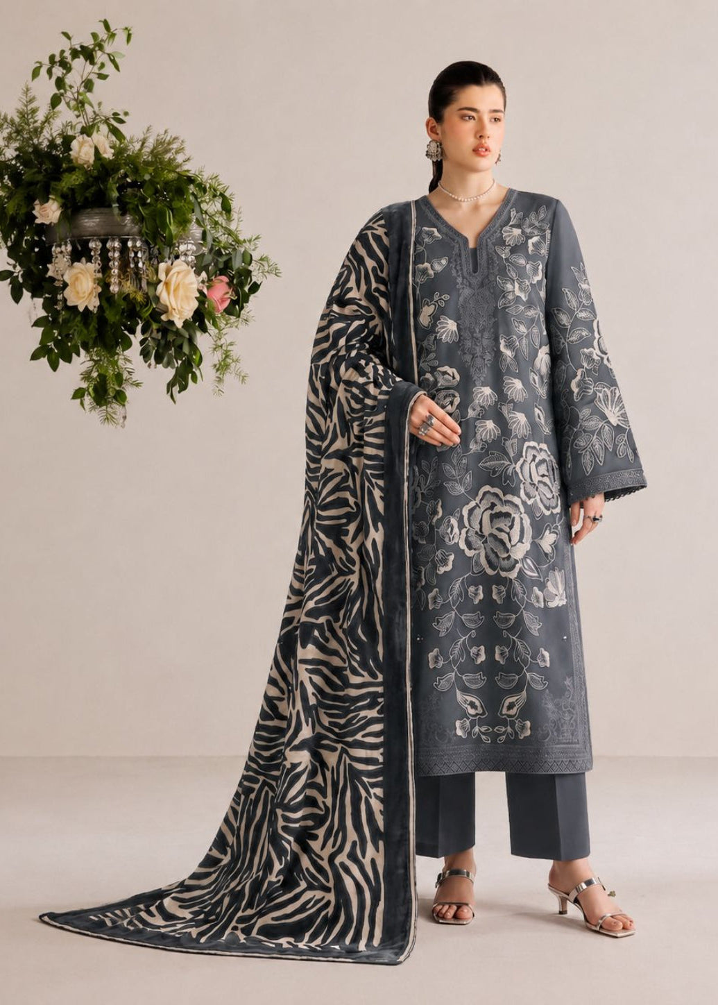 A woman models a three-piece traditional outfit consisting of a dark gray embroidered kurta, matching trousers, and a long printed dupatta with a zebra-like pattern. She is standing in a studio setting with a hanging floral arrangement on the left.