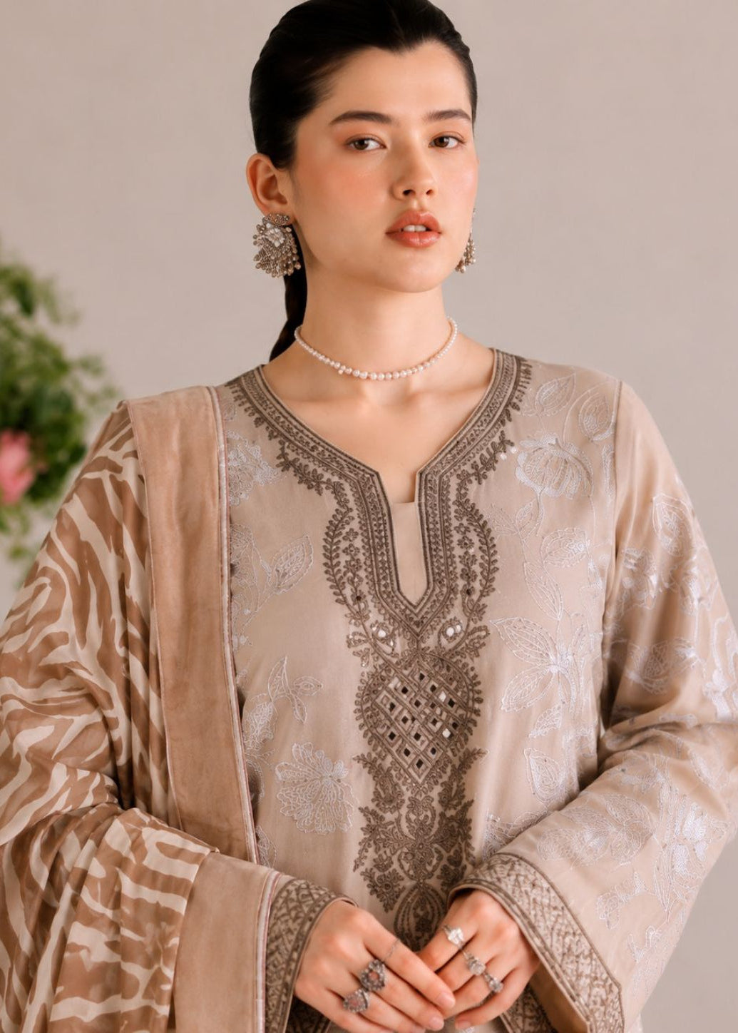 A woman models a traditional beige and brown embroidered ethnic suit with a matching patterned dupatta, set against a soft neutral background with a blurred floral arrangement.