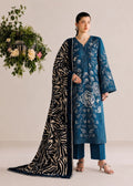A woman models a traditional blue embroidered kurta set with matching pants and a black-and-beige patterned dupatta, posed against a neutral beige studio background with a decorative floral arrangement.