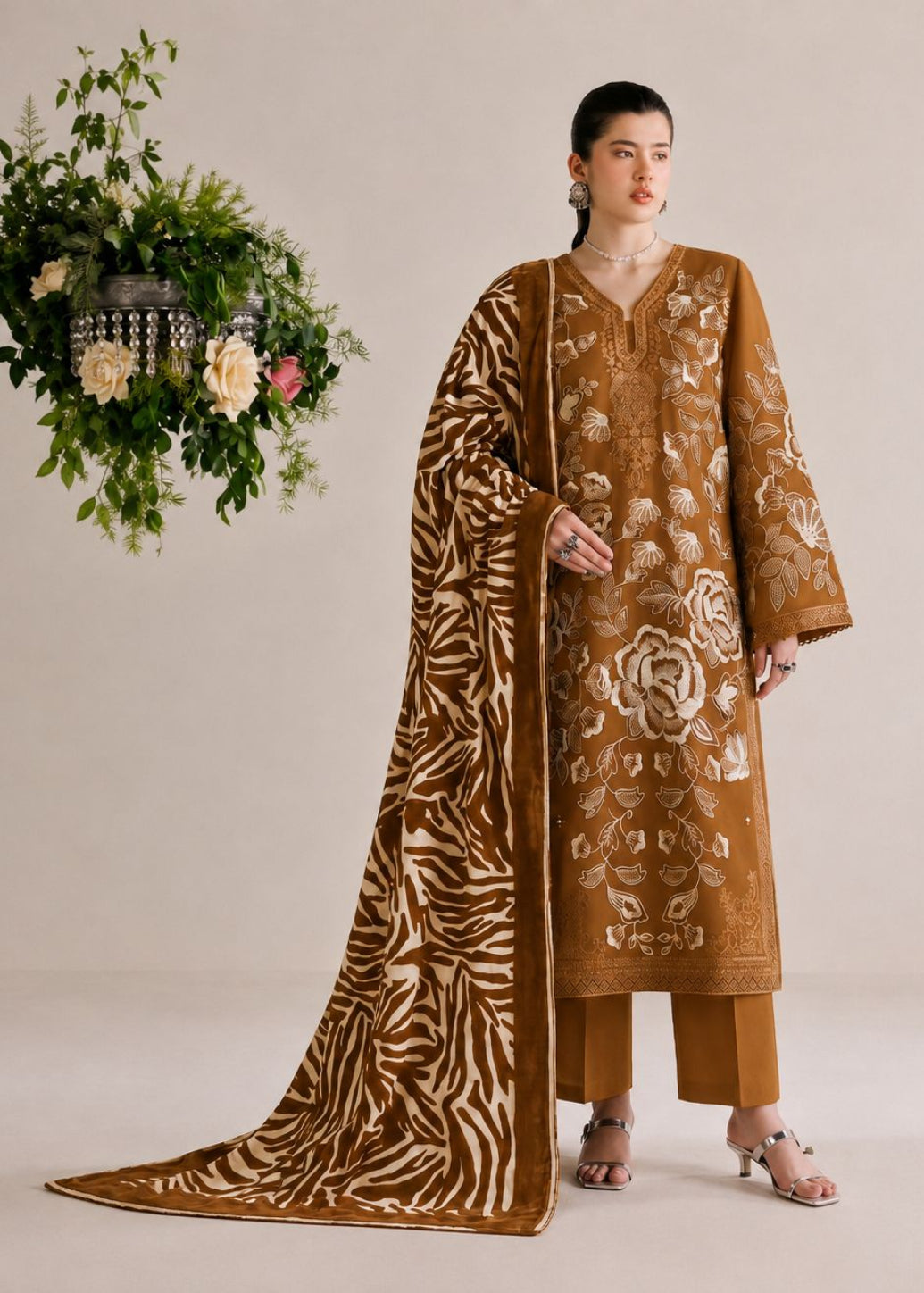 A woman models a traditional brown salwar kameez suit with intricate gold floral embroidery and a matching patterned dupatta, set against a neutral beige background with a hanging floral arrangement.