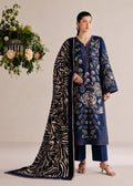A model stands in a studio wearing a navy blue traditional outfit with intricate gold floral embroidery, matching navy pants, and a long black dupatta with gold zari borders and motifs. A hanging floral arrangement is on the left side against a plain beige background.