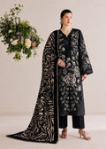 A woman models a black three-piece traditional outfit with beige floral embroidery, a matching black dupatta with beige zebra-like stripes, and black trousers, set against a warm beige studio background with a hanging floral arrangement.