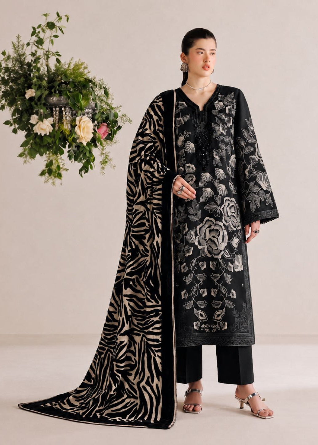 A woman models a black three-piece traditional outfit with beige floral embroidery, a matching black dupatta with beige zebra-like stripes, and black trousers, set against a warm beige studio background with a hanging floral arrangement.