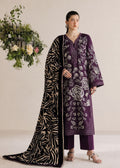 A model stands in a studio wearing a deep purple traditional outfit with white floral embroidery, matching purple pants, and a black-and-white patterned dupatta. A large floral arrangement hangs on the left against a plain beige background.